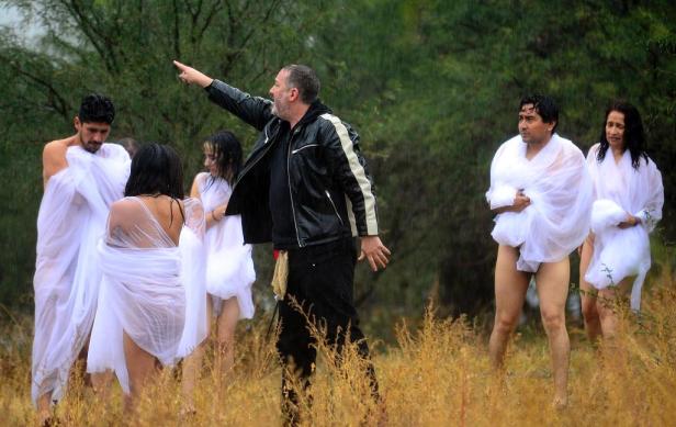 Eine Gruppe von Menschen, nur mit weißen Tüchern bekleidet, steht im Regen auf einem Feld. Ein Mann in Jacke zeigt mit dem Finger.