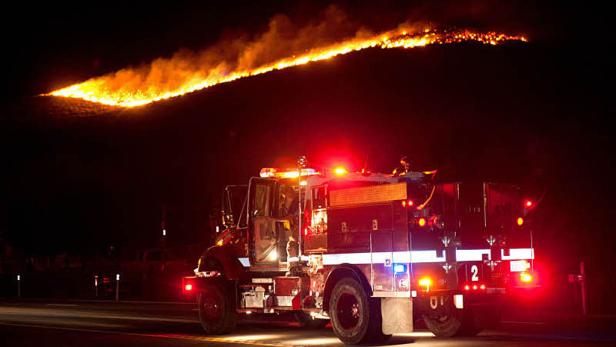 Ein Feuerwehrwagen steht nachts vor einem Hügel, der von einem Waldbrand betroffen ist.
