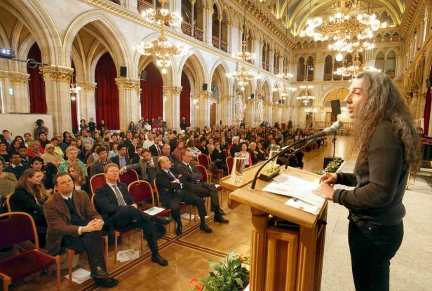 Eine Rednerin spricht vor einem großen Publikum in einem prunkvollen Saal.