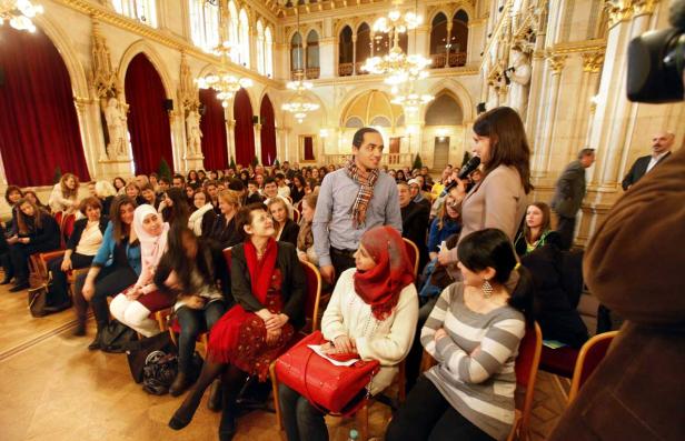 Eine Frau spricht bei einer Veranstaltung vor einem Publikum in einem verzierten Saal.