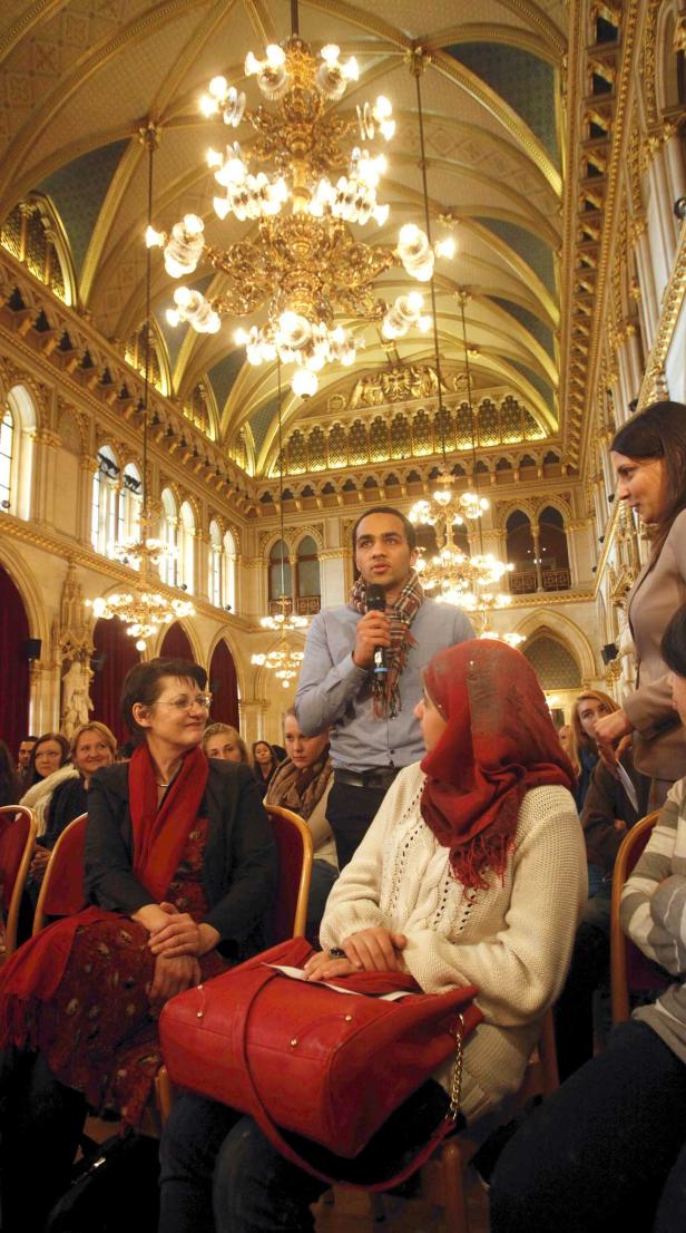 Ein Mann spricht in einem reich verzierten Saal mit Kronleuchtern zu einem Publikum.
