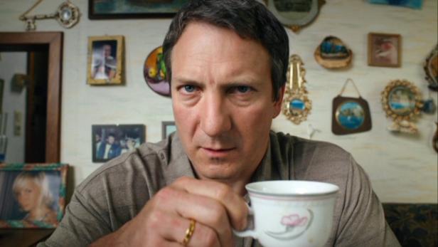 Ein Mann hält eine Teetasse in der Hand, im Hintergrund eine Wand voller Bilder.