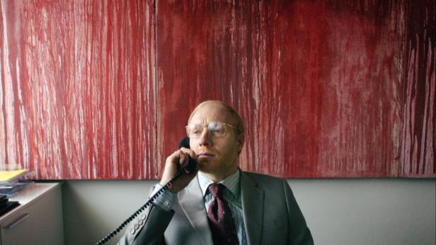 Ein Mann mit Brille telefoniert vor einem großen, roten Gemälde.