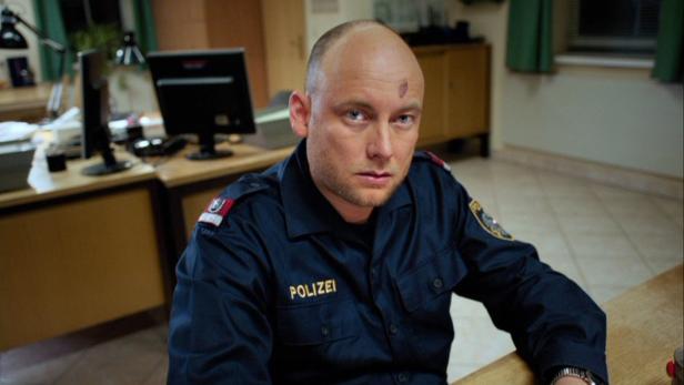 Ein Polizist mit einer Schramme auf der Stirn sitzt an einem Schreibtisch.