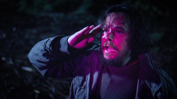 Ein Mann mit Bart und Jacke salutiert im Dunkeln.