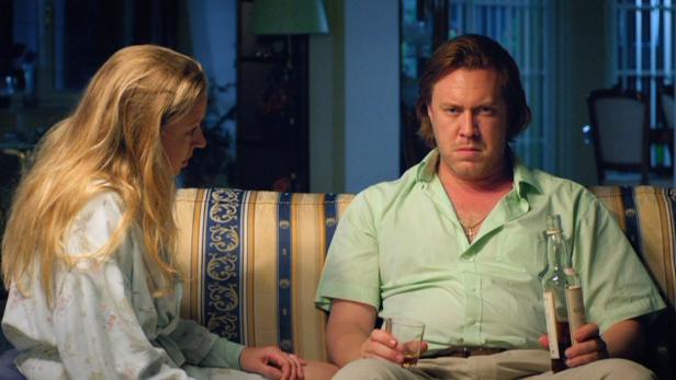 Ein Mann sitzt mit einer Flasche und einem Glas auf einem Sofa neben einer Frau.