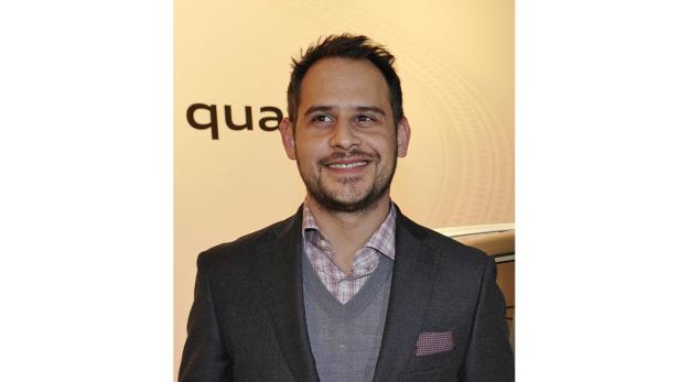 Ein lächelnder Mann in einem grauen Anzug vor einem hellen Hintergrund.