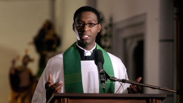 Ein schwarzer Pfarrer hält eine Rede an einem Rednerpult.