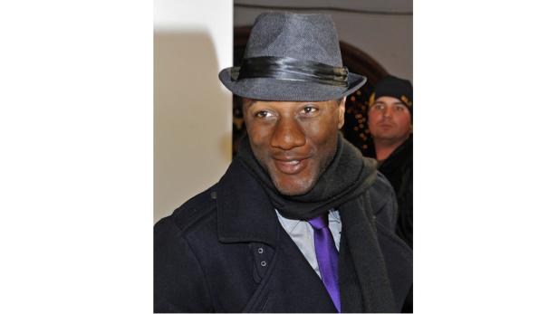 Ein Mann mit Hut, Schal und Mantel lächelt in die Kamera.