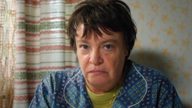 Eine Frau mit kurzem, dunklem Haar und blauem Pyjama-Oberteil blickt ernst in die Kamera.