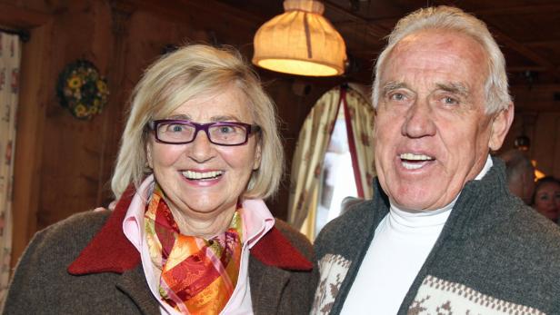 Ein älteres Paar lächelt in die Kamera, die Frau mit Brille und buntem Tuch, der Mann mit Strickjacke.