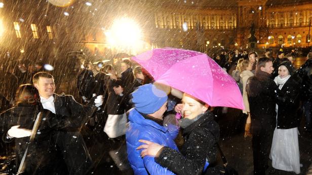 Trotz Schneefalls tanzen Menschen auf einem Platz, einige unter einem pinkfarbenen Regenschirm.