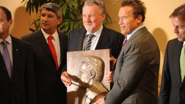 Arnold Schwarzenegger hält eine Bronzebüste von sich selbst mit anderen Männern im Anzug.