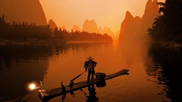 Ein Fischer stakt sein Bambusfloß auf einem ruhigen Fluss entlang, im Hintergrund ragen Berge auf.