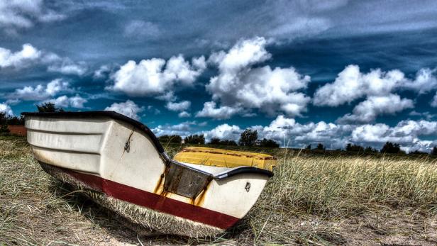 Ein weißes Ruderboot liegt auf einer sandigen Wiese unter einem bewölkten Himmel.