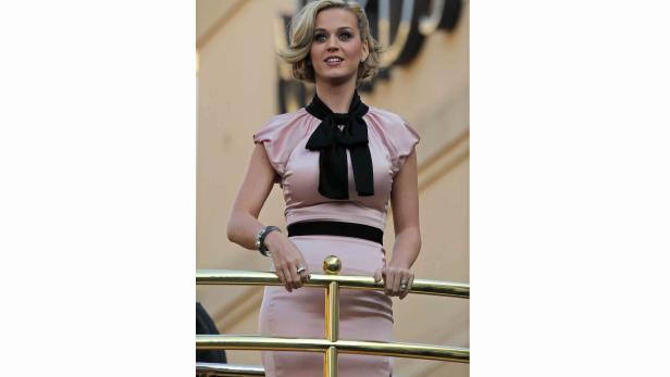 Katy Perry in einem rosa Kleid mit schwarzer Schleife an einem goldenen Geländer.