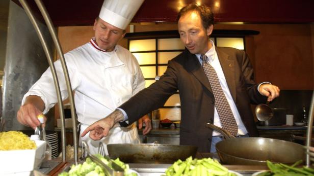 Ein Koch und ein Mann im Anzug stehen in einer Küche und bereiten Speisen zu.