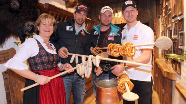 Eine Frau im Dirndl und drei Männer präsentieren Würste und Brezeln auf Holzstäben.