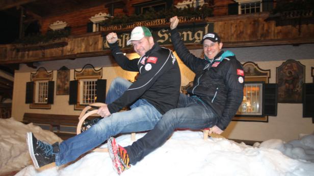 Zwei Männer sitzen auf einem Holzschlitten im Schnee und jubeln.