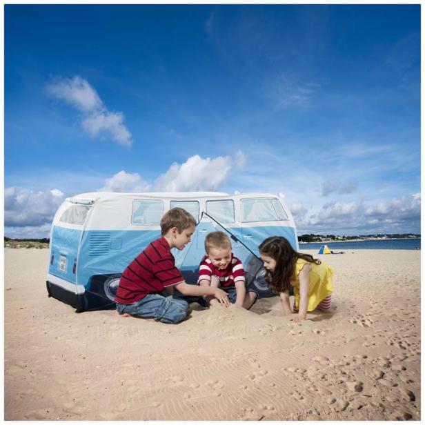 Drei Kinder spielen am Strand vor einem VW-Bus-Zelt.