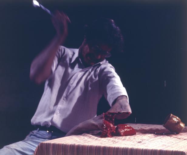 Ein Mann hackt mit einem Messer auf einen abgetrennten Arm auf einem Tisch.