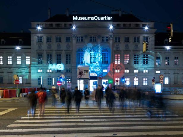 Das MuseumsQuartier in Wien ist nachts beleuchtet, Menschen überqueren einen Zebrastreifen.