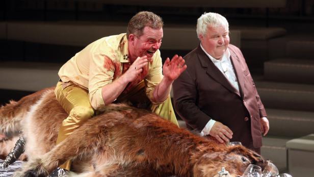 Ein Mann, mit Blut bedeckt, reitet auf einem Tierfell, während ein anderer Mann daneben steht.