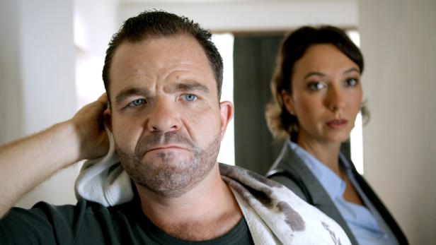 Ein Mann mit einem Handtuch um den Hals und eine Frau im Hintergrund.