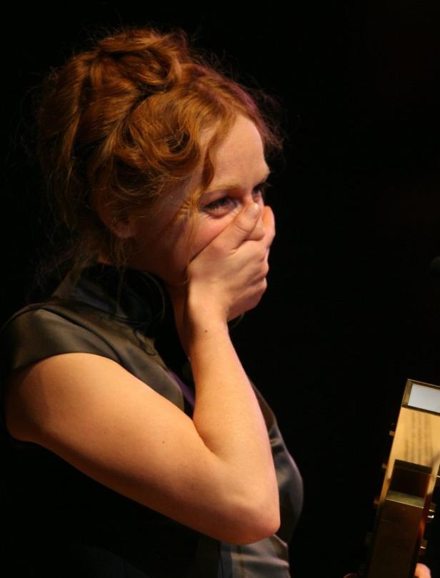 Eine Frau mit roten Haaren hält sich vor gerührter Emotion die Hand vor den Mund.