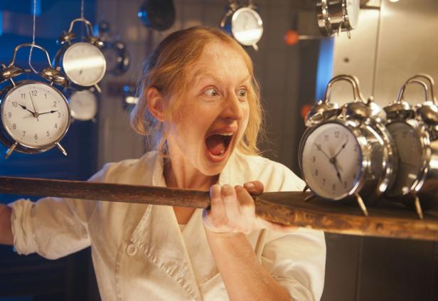 Eine Frau in Kochkleidung schreit entsetzt, während um sie herum viele Wecker hängen.