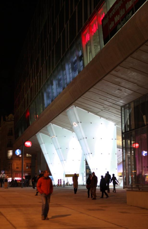 Passanten vor einem modernen Gebäude mit beleuchteten Glasstützen in der Nacht.