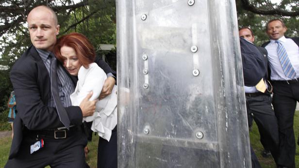 Ein Mann schützt Julia Gillard mit einem Schutzschild vor Demonstranten.