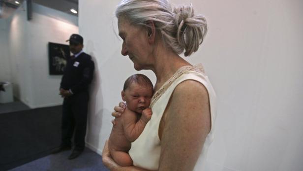 Eine ältere Frau hält eine lebensechte Babypuppe im Arm, im Hintergrund ein Wachmann.