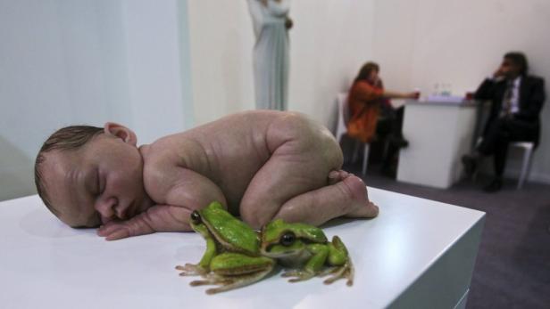 Ein schlafendes Baby liegt neben zwei grünen Fröschen auf einer weißen Oberfläche.