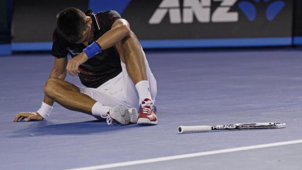 Novak Đoković sitzt erschöpft auf dem Tennisplatz, sein Schläger liegt daneben.