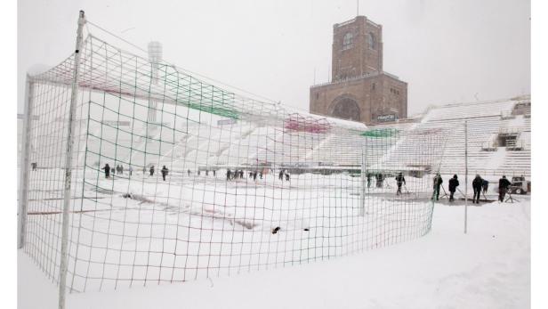 Ein verschneites Fußballstadion mit Arbeitern, die den Schnee räumen.