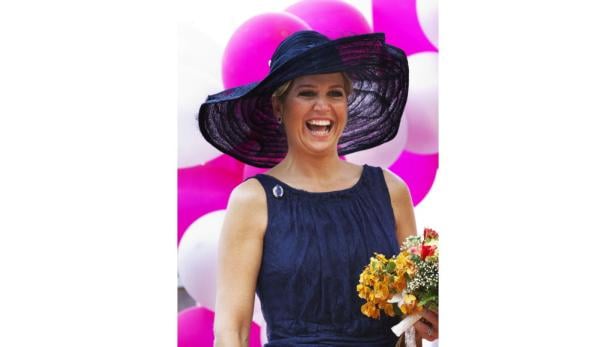 Königin Máxima lacht mit einem Blumenstrauß und einem großen Hut vor einem Hintergrund aus Ballons.
