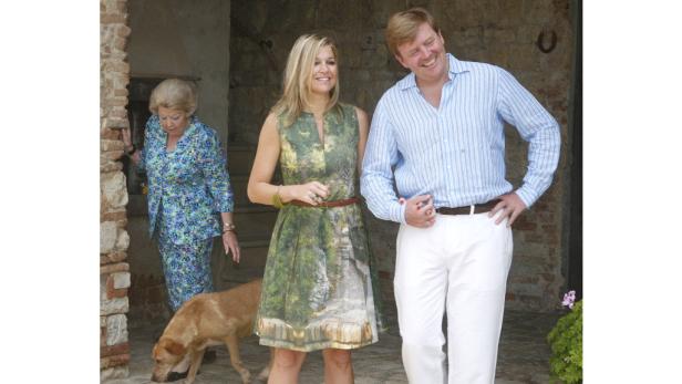 Königin Beatrix, Königin Máxima und König Willem-Alexander posieren mit einem Hund vor einem Gebäude.