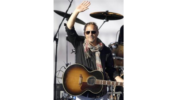 Kevin Costner mit Gitarre auf der Bühne winkt dem Publikum zu.