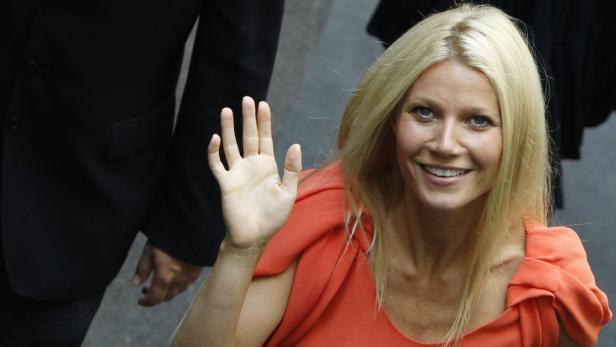 Gwyneth Paltrow lächelt und winkt in einem orangefarbenen Kleid.