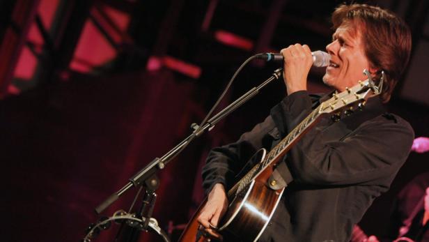 Kevin Bacon singt und spielt Gitarre auf einer Bühne.