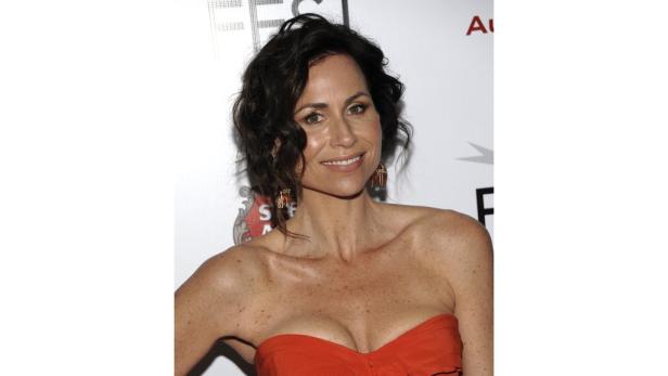 Minnie Driver in einem roten, trägerlosen Kleid lächelt in die Kamera.