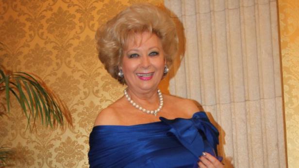 Eine ältere Frau mit blonden Haaren und einem blauen Kleid lächelt in die Kamera.