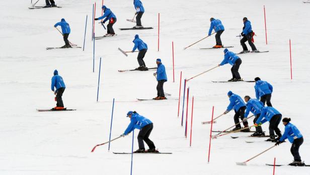 Eine Gruppe von Skifahrern präpariert eine Skipiste mit Rechen zwischen roten und blauen Stangen.