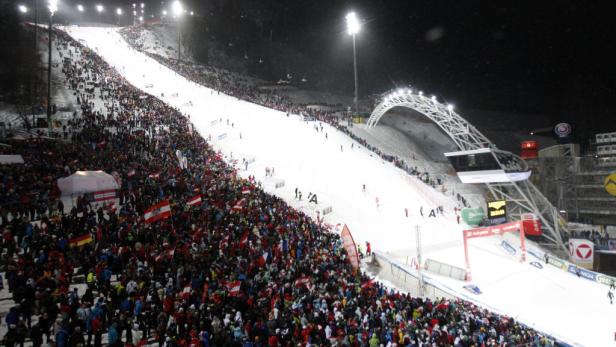 Eine große Menschenmenge mit österreichischen Flaggen säumt eine Skipiste bei Nacht.