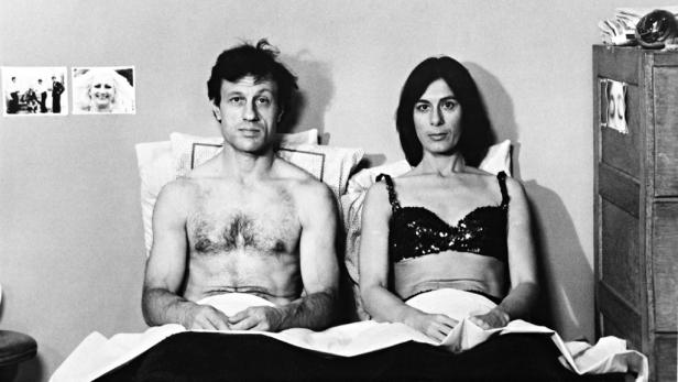 Ein Mann und eine Frau sitzen in einem Bett, beide mit ernstem Gesichtsausdruck.