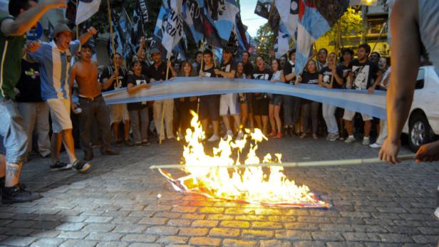 Eine Gruppe von Menschen hält Flaggen und ein Banner, während eine Flagge am Boden brennt.