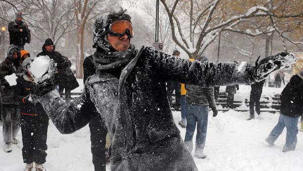 Eine Person mit Skibrille wirft einen Schneeball in einer verschneiten Parklandschaft.