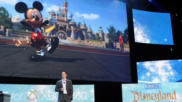 Ein Mann präsentiert das Spiel „Kinect Disneyland Adventures“ mit Micky Maus im Hintergrund.