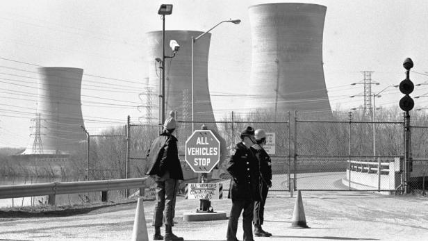 Drei Personen stehen vor dem Eingang zu einem Atomkraftwerk, im Hintergrund Kühltürme.